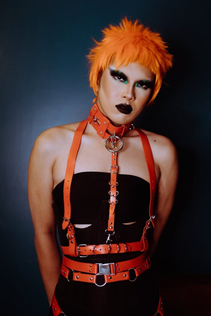 Self assured young ethnic androgynous male model with makeup in orange wig and BDSM collar and harness looking at camera against blue background